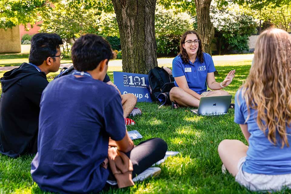 A female SLU 101 leader speaks to a group of students. The group is sitting on the grass on a sunny day.