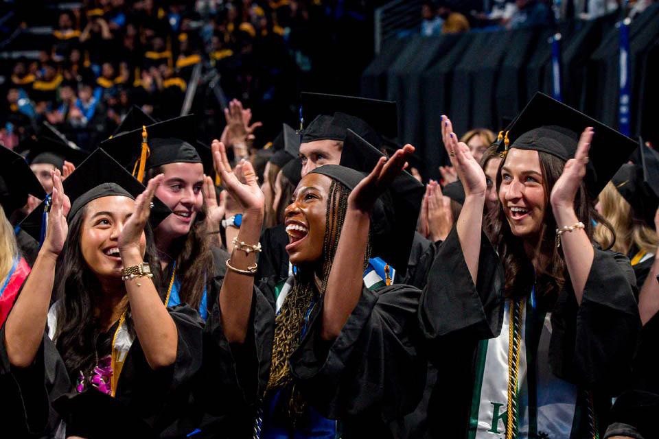 Students cheer and clap wearing graduation gowns.