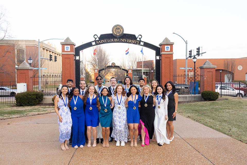 A large group of students dressed nicely and with medals around their neck pose for a photo outside on SLU's campus.