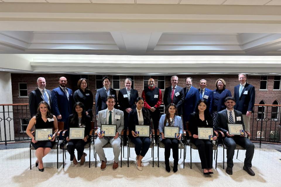 2024 Award Winners A group of students sit while holding awards plaques while professors and supporters stand behind them.