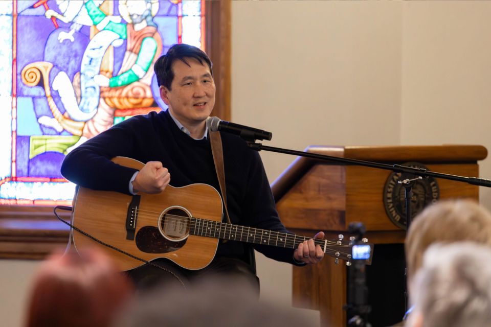 James Rhee Speaker Series 2026 a man sits with a guitar speaking in front of a crowd of people
