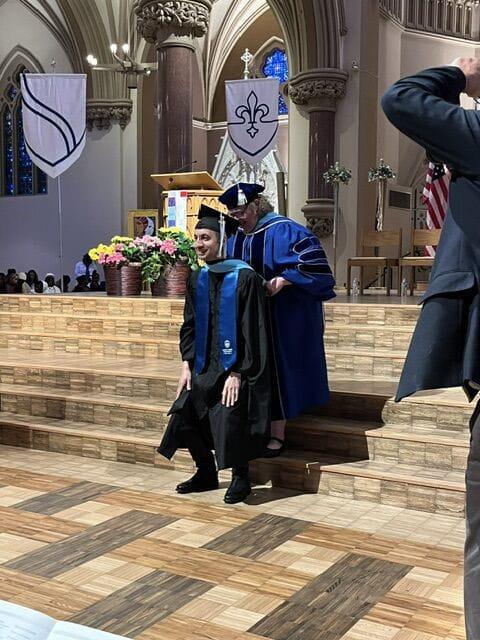 A graduate in cap and gown stands in front of a church where a professor places an academic hood around his neck.
