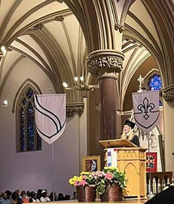 A speaker in a graduation cap and gown speaks at a pulpit at the front of a church.