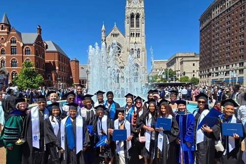 A group of graduates in caps and gowns pose for a photo in front of a fountain outside of St. Francis Xavier Church
