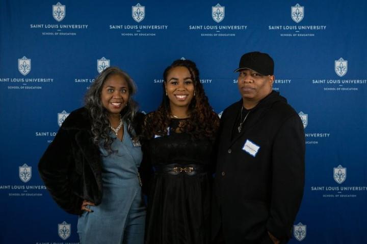 Three people smile at the camera while standing in front of a Saint Louis University backdrop