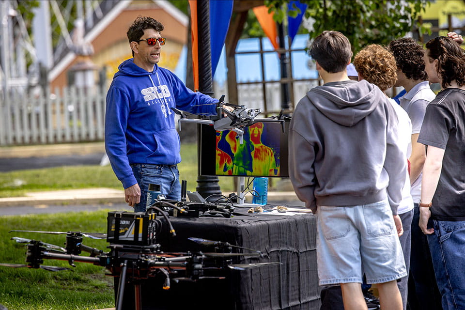A student wearing a SLU sweatshirt demonstrates equipment including a video screen and a drone to a group of students.tes geospatial equipment including a 