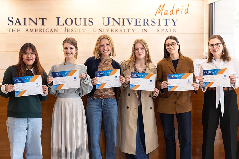 Six women hold certificates and smile for photo with the SLU-Madrid logo in the background.