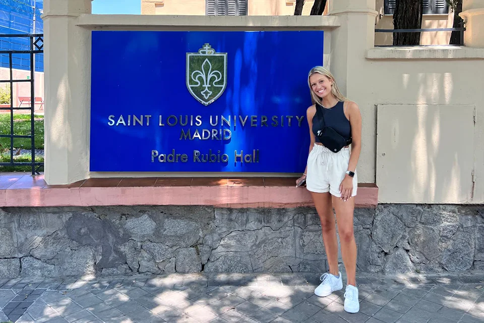 Emily Risner stands smiling beside the Saint Louis University-Madrid sign outside Padre Rubio Hall. She is wearing a black sleeveless top, white shorts and white sneakers, with trees and the campus entrance visible in the background.
