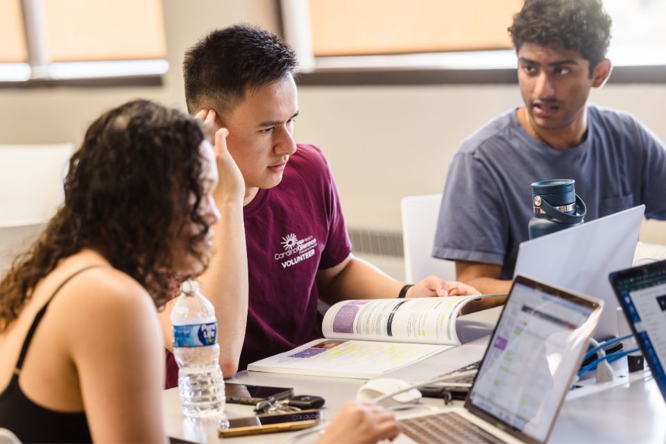Three students site at a table with open textbooks and laptops.