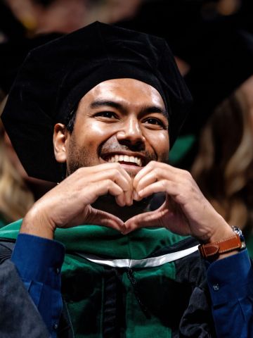 A medical graduate in a cap and gown smiles and holds his hands in the shape of a heart.