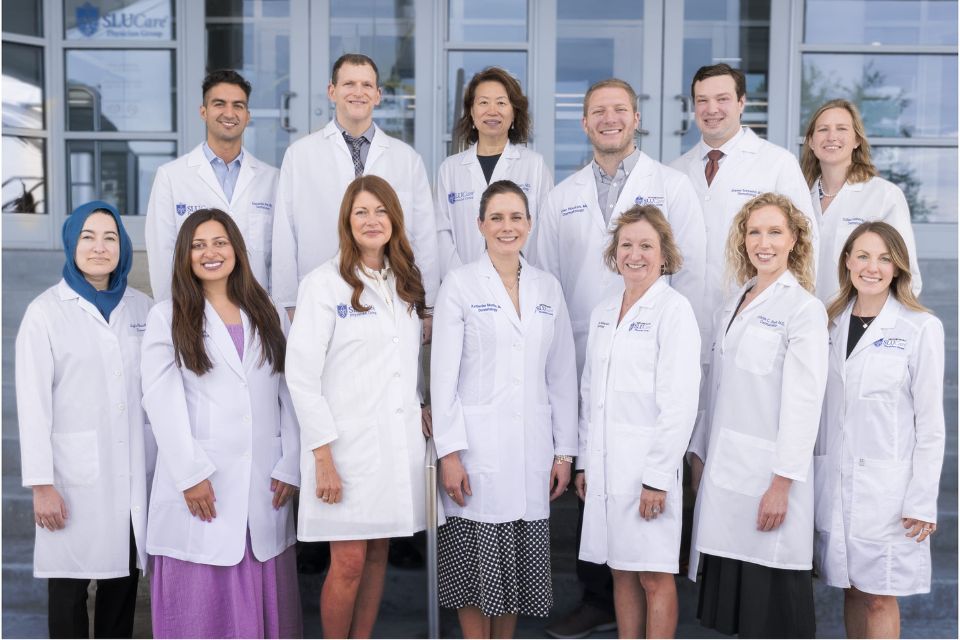 A group of doctors in white coats smile and pose together outside an office building entrance.