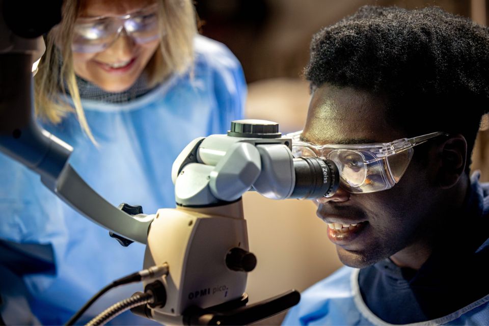 An instructor stands beside a student in protective goggles looking through a microscope.