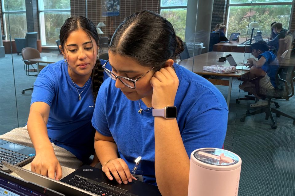Two students look closely at a laptop together while working at a desk inside a university library.