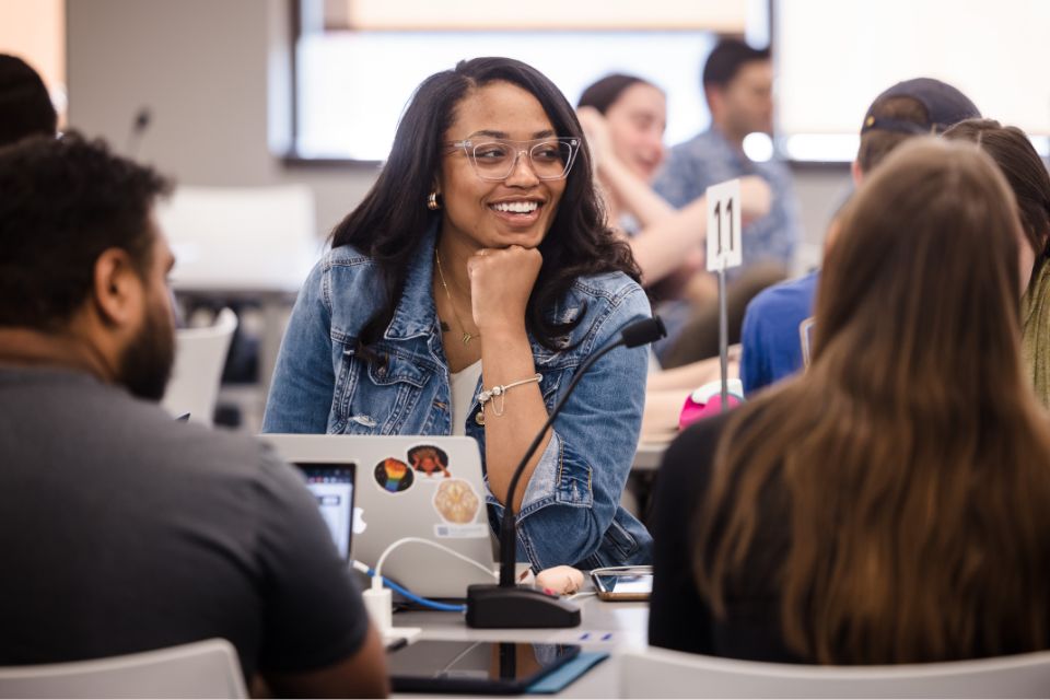 A student sits at a conference table with an open laptop, talking during class.