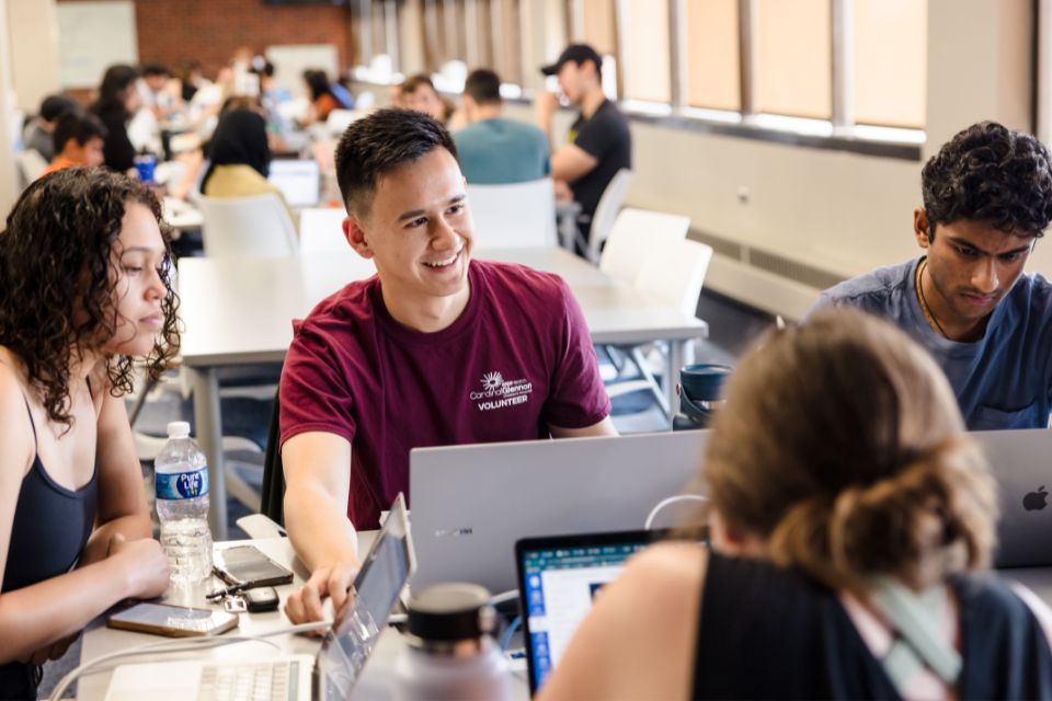 A diverse group of students sit together at a table in a collaborative workspace, smiling and working on laptops.