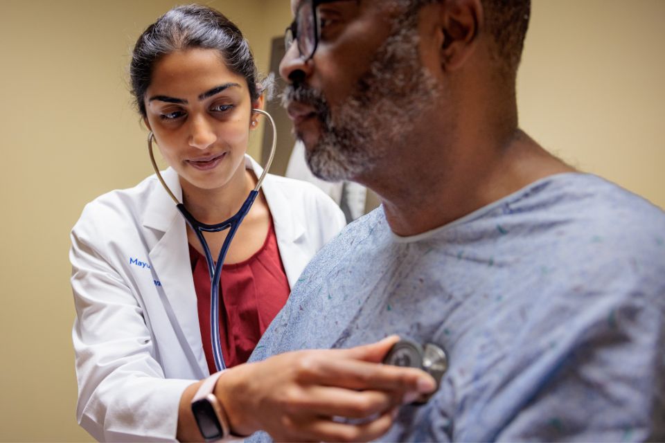A medical student in a white coat uses a stethoscope to examine a sitting patient.