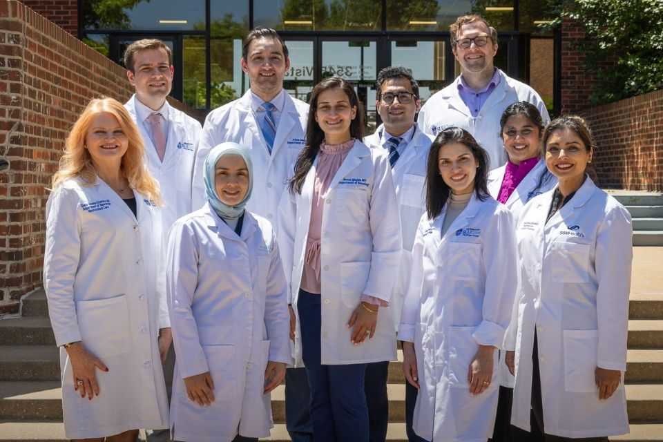 A group photo of 2026 neurology graduates in white coats standing outside a brick building entrance