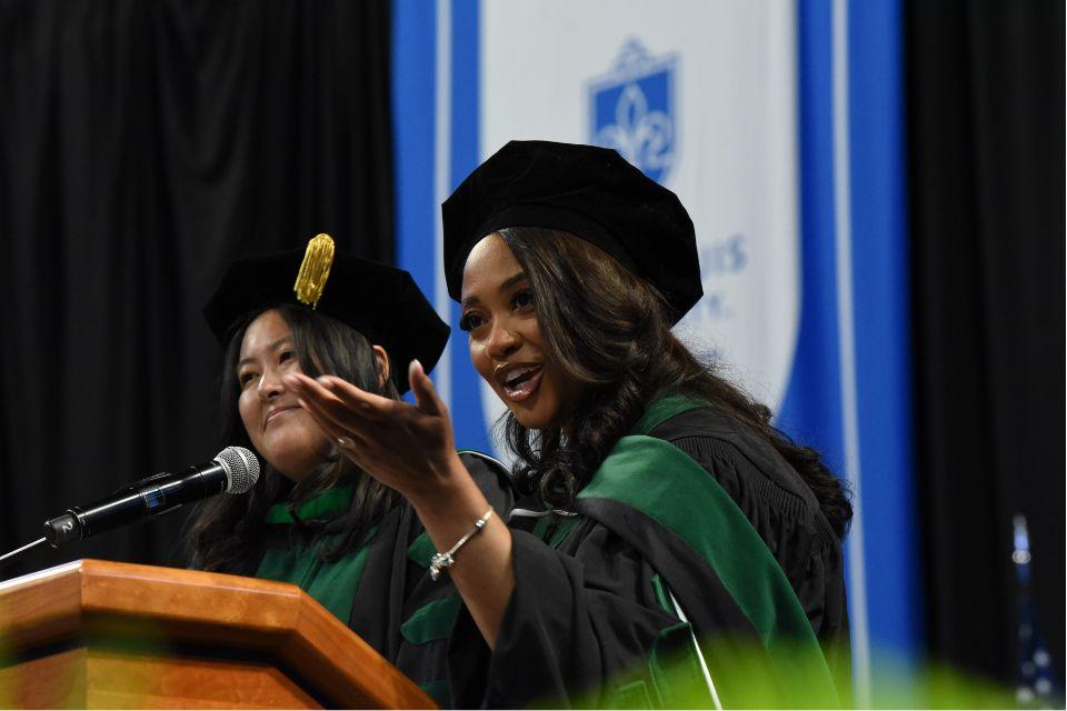 Two students dressed in caps and gowns stand at a podium in front of a blue banner and to address an audience.