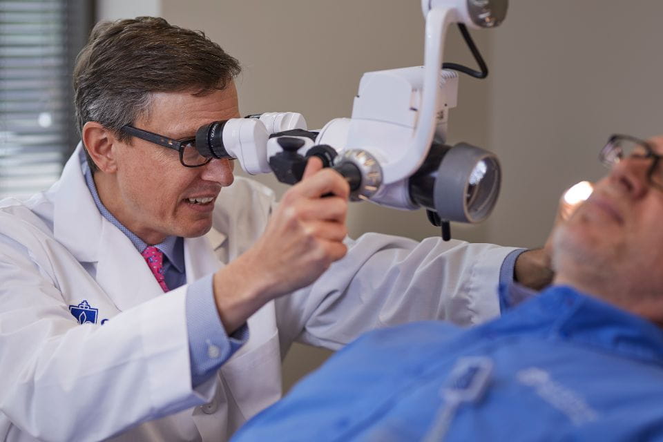 Inside a hospital room, an otolaryngologist peers through a lens to examine a patient.
