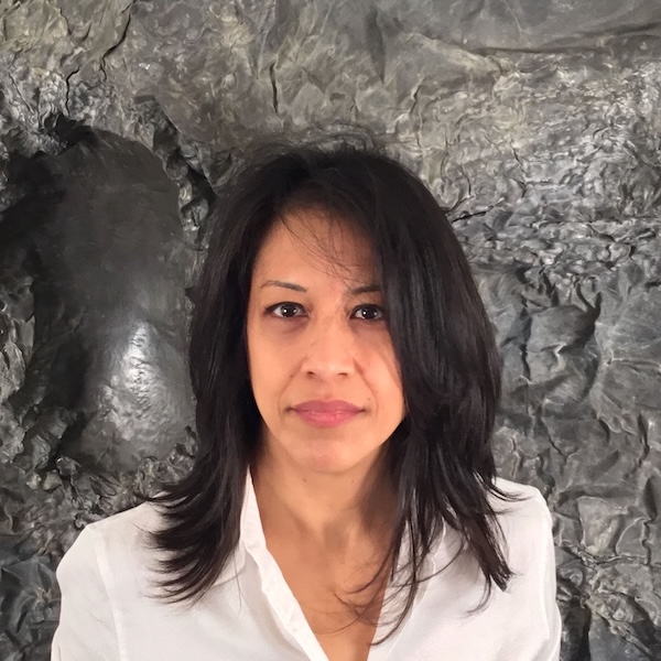 Athena LaTocha A woman with shoulder-length black hair wearing a white shirt looks directly at the viewer. Visible from the shoulders up, she stands in front of a textured slate gray background