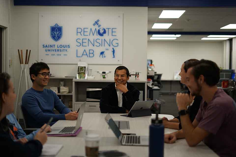 Geospatial Environment Five people sit around a table while working on their laptops. A white and blue sign in the background reads Saint Louis University Remote Sensing Lab.
